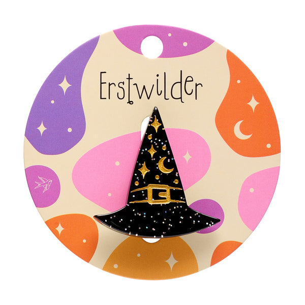 A glitter black witch hat acrylic resin brooch decorated with gold glitter stars, a crescent moon, and a gold buckle, mounted on a colorful round backing card with purple, orange, and pink abstract shapes.