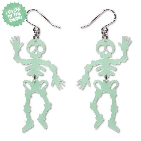 A pair of dangling skeleton-shaped earrings in a pale mint green glow-in-the-dark finish, shown on a plain white background with a green starburst sticker that reads 'I GLOW IN THE DARK!'.