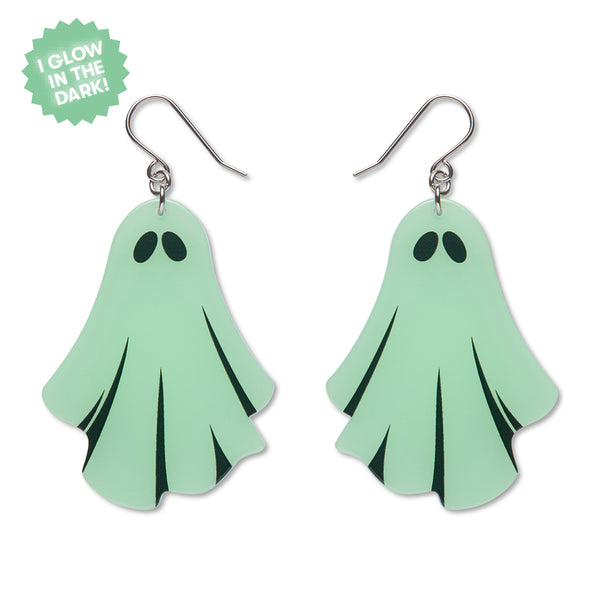 A pair of dangle earrings shaped like cute ghost figures in pale mint green, with black eyes and black shadow lines. A green starburst sticker in the top left corner reads 'I GLOW IN THE DARK!'.