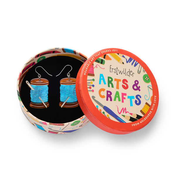 pair of spools of blue thread with metallic silver needle acrylic resin dangle earrings, shown in illustrated round box packaging