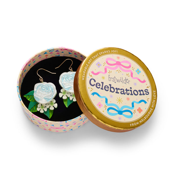A pair of dangling earrings featuring light blue and white rose charms, green leaf accents, and small white flowers, suspended from gold-tone hooks, shown in illustrated round gift box packaging.