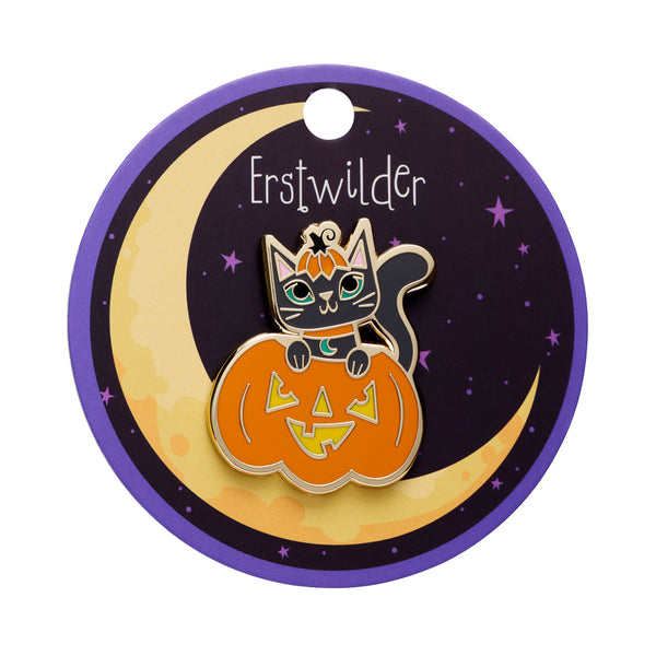 A gold metal enamel pin featuring a cartoon black cat wearing an orange pumpkin hat and sitting in a jack-o'-lantern, mounted on a round backing card with a purple border and the text 'Erstwilder'.