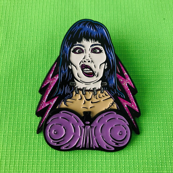 matte finish black metal enamel pin of Elizabeth Shelley from the movie Frankenhooker in front of glittery pink lightning bolt background with articulated purple bra detail. Shown on neon green fabric background