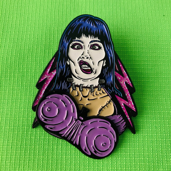 matte finish black metal enamel pin of Elizabeth Shelley from the movie Frankenhooker in front of glittery pink lightning bolt background with articulated purple bra detail. Shown on neon green fabric background with bra moved to the right