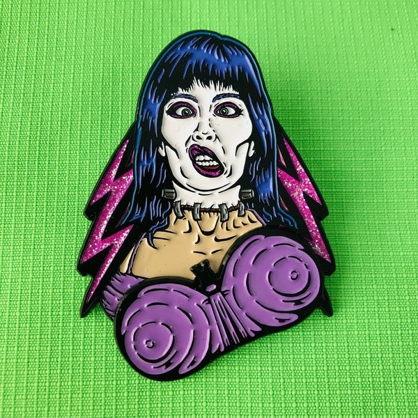 matte finish black metal enamel pin of Elizabeth Shelley from the movie Frankenhooker in front of glittery pink lightning bolt background with articulated purple bra detail. Shown on neon green fabric background with bra moved to the left