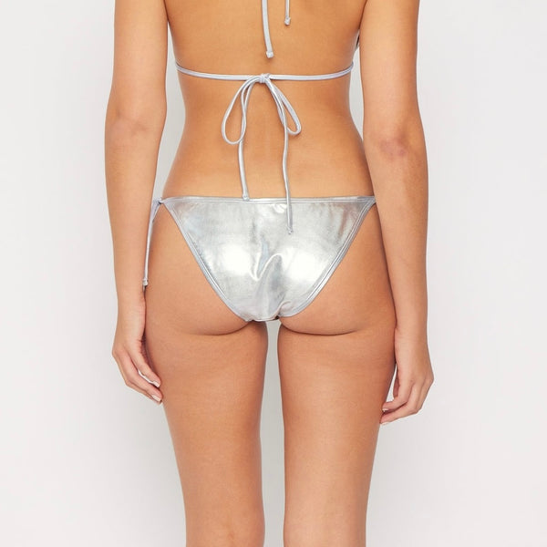 Model wearing low-rise string bikini bottom made of silver stretch lamé material. Seen from the back