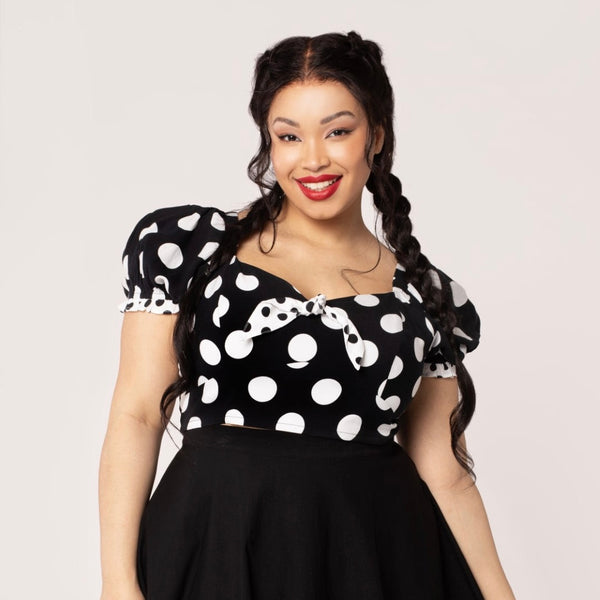 A plus size model wearing a short sleeve crop top with puff sleeves and a sweetheart neckline. It has a pattern of large white on black polka dots with small contrasting black on white dots at the cuffs and knotted tie detail at the bodice. Shown from the front