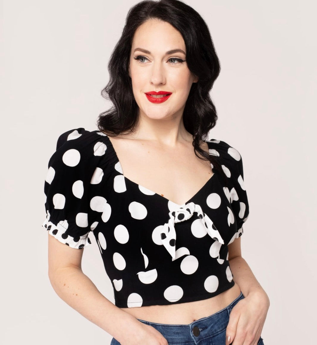 A model wearing a short sleeve crop top with puff sleeves and a sweetheart neckline. It has a pattern of large white on black polka dots with small contrasting black on white dots at the cuffs and knotted tie detail at the bodice. Shown from the front 