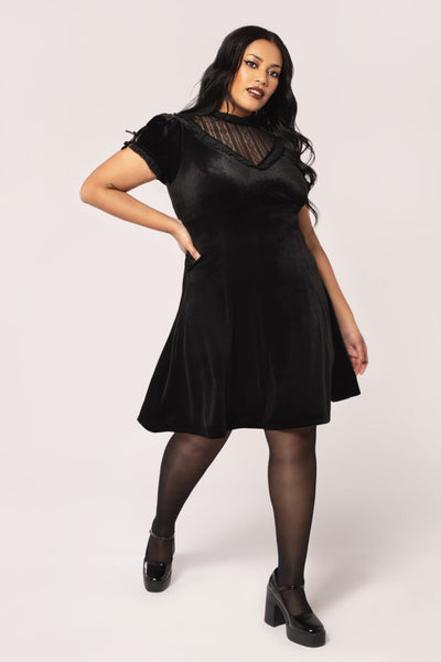 Plus sized model wearing a black velvet mini dress with high frilled neckline, V-shaped panel of floral striped lace at the bodice, slightly puffed sleeves with lace trim & keyhole detail with black ribbon bows. Skirt ends above the knee. Shown from front