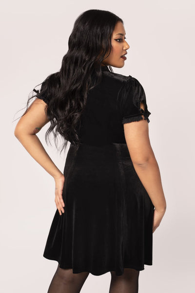 Plus sized model wearing a black velvet mini dress with high frilled neckline, V-shaped panel of floral striped lace at the bodice, slightly puffed sleeves with lace trim & keyhole detail with black ribbon bows. Skirt ends above the knee. Shown from back to display keyhole