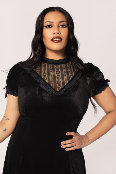 Plus sized model wearing a black velvet mini dress with high frilled neckline, V-shaped panel of floral striped lace at the bodice, slightly puffed sleeves with lace trim & keyhole detail with black ribbon bows. Skirt ends above the knee. Shown from front in close up of bodice