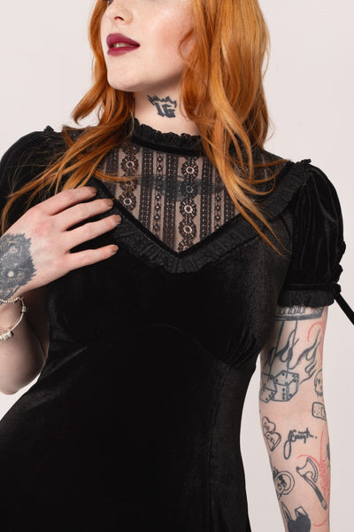 Model wearing a black velvet mini dress with high frilled neckline, V-shaped panel of floral striped lace at the bodice, slightly puffed sleeves with lace trim & keyhole detail with black ribbon bows. Skirt ends above the knee. Shown in close up of bodice