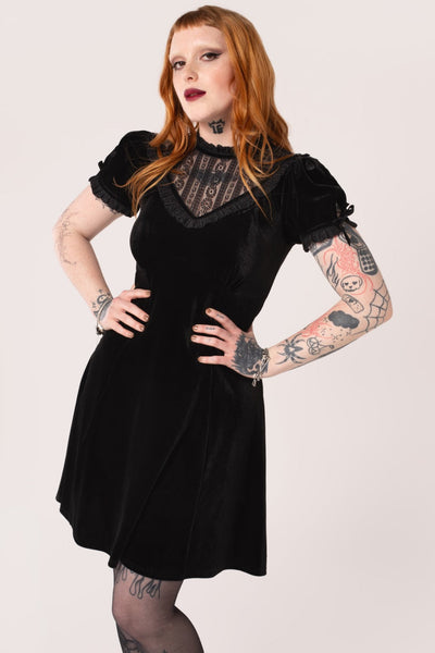 Model wearing a black velvet mini dress with high frilled neckline, V-shaped panel of floral striped lace at the bodice, slightly puffed sleeves with lace trim & keyhole detail with black ribbon bows. Skirt ends above the knee. Shown from front