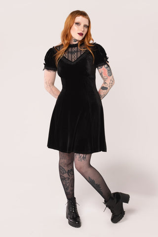 Model wearing a black velvet mini dress with high frilled neckline, V-shaped panel of floral striped lace at the bodice, slightly puffed sleeves with lace trim & keyhole detail with black ribbon bows. Skirt ends above the knee. Shown from front