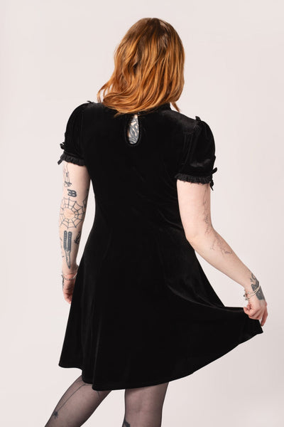 Model wearing a black velvet mini dress with high frilled neckline, V-shaped panel of floral striped lace at the bodice, slightly puffed sleeves with lace trim & keyhole detail with black ribbon bows. Skirt ends above the knee. Shown from back to display keyhole