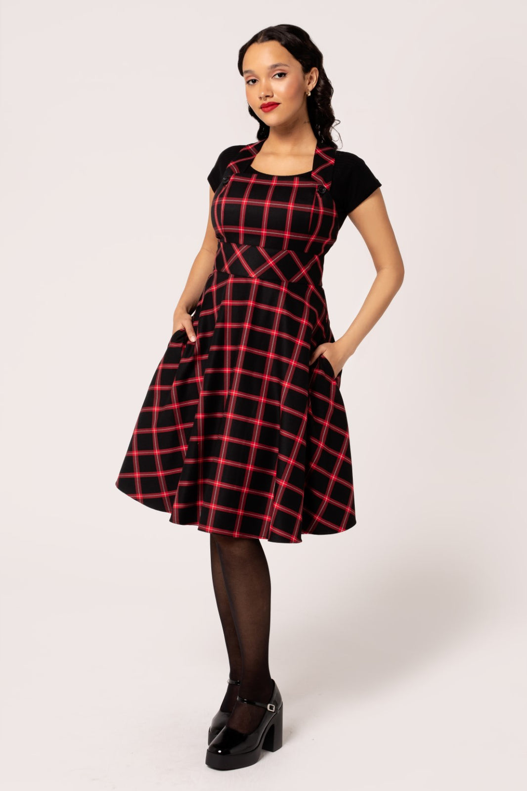 Model wearing black and red plaid pinafore style dress with wide adjustable straps with black plastic buttons, a like wide waistband, and a full knee length skirt with side seam pockets. Shown from the front