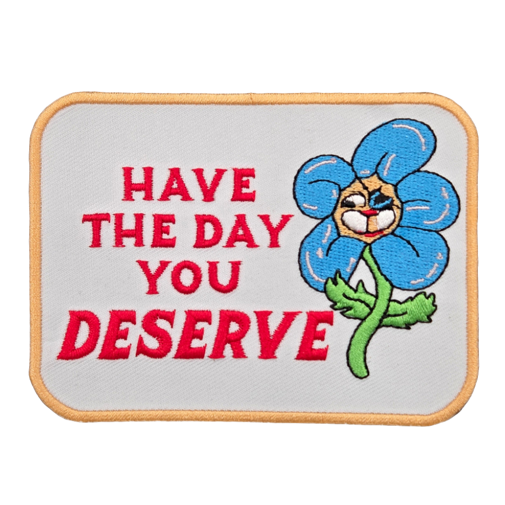rectangular white embroidered patch with yellow border and green & blue daisy with funny face alongside "HAVE THE DAY YOU DESERVE" written in red script
