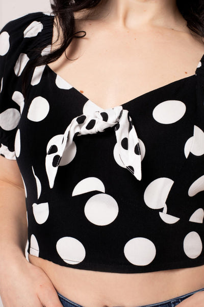 A model wearing a short sleeve crop top with puff sleeves and a sweetheart neckline. It has a pattern of large white on black polka dots with small contrasting black on white dots at the cuffs and knotted tie detail at the bodice. Shown from the front in close up