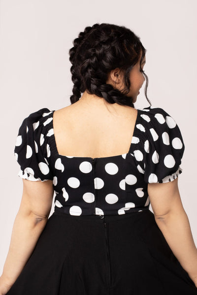 A plus size model wearing a short sleeve crop top with puff sleeves and a sweetheart neckline. It has a pattern of large white on black polka dots with small contrasting black on white dots at the cuffs and knotted tie detail at the bodice. Shown from the back