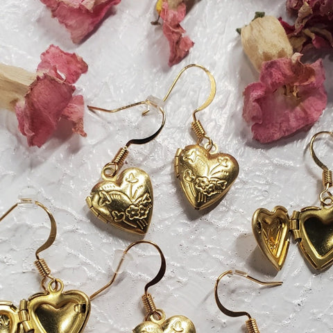Several pairs of gold dangle earrings with locket heart charms, shown open and closed on a white marbled background with rose petals