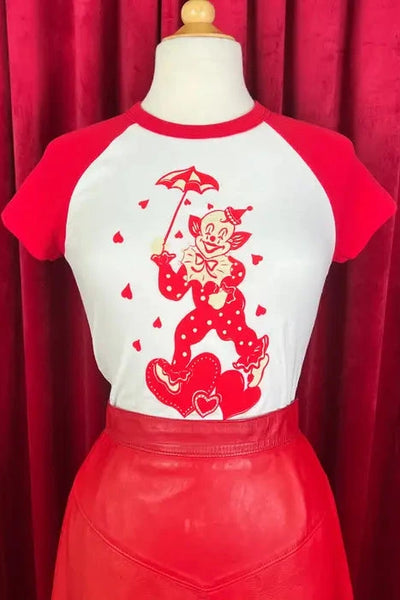 Short sleeved crop white t-shirt with red raglan sleeves and red & cream illustration of a clown wearing a polka dotted outfit holding an umbrella standing on a trio of hearts and surrounded by hearts raining down. Shown tucked into a red skirt on a dress form