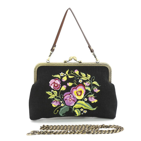 vintage-inspired handbag with an antiqued bronze metal kiss lock closure featuring purple and yellow pansies in bloom surrounded by buds and leaves embroidered on a black fabric background. Comes with a removable brown faux leather handbag strap and antiqued bronze metal curb link shoulder strap. Strap is shown in front of bag with hand strap attached 