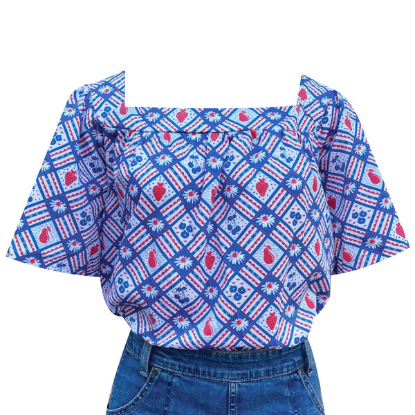 A square necked and bell sleeved peasant style top with an all-over checkered pattern of daisies, strawberries, cherries, blueberries, and pears. Seen tucked into a denim skirt