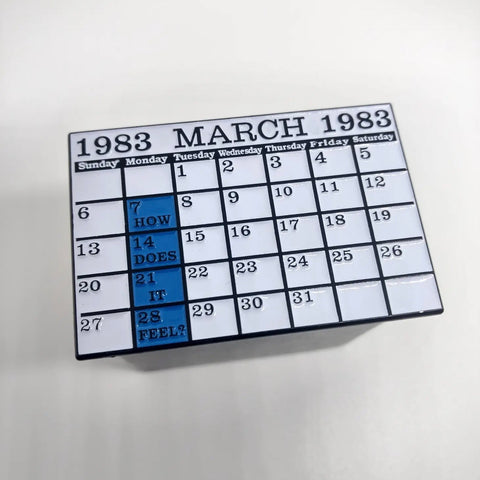 A rectangular enamel pin in the shape of a calendar with the date March 1983. Each Monday spot is colored blue with the words “How does it feel” under each date while the rest of the calendar is white