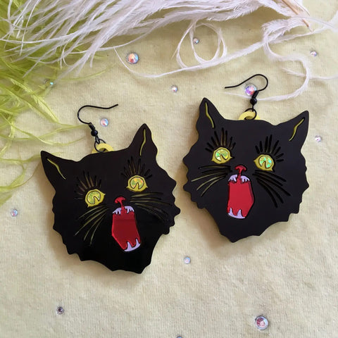 Dangle earrings in the shape of  pair of black cats reminiscent of vintage Halloween decorations crafted out of layered laser-cut acrylic: black, mirrored red mouth, pearly iridescent teeth and yellow glitter foil eyes with a solid neon yellow back