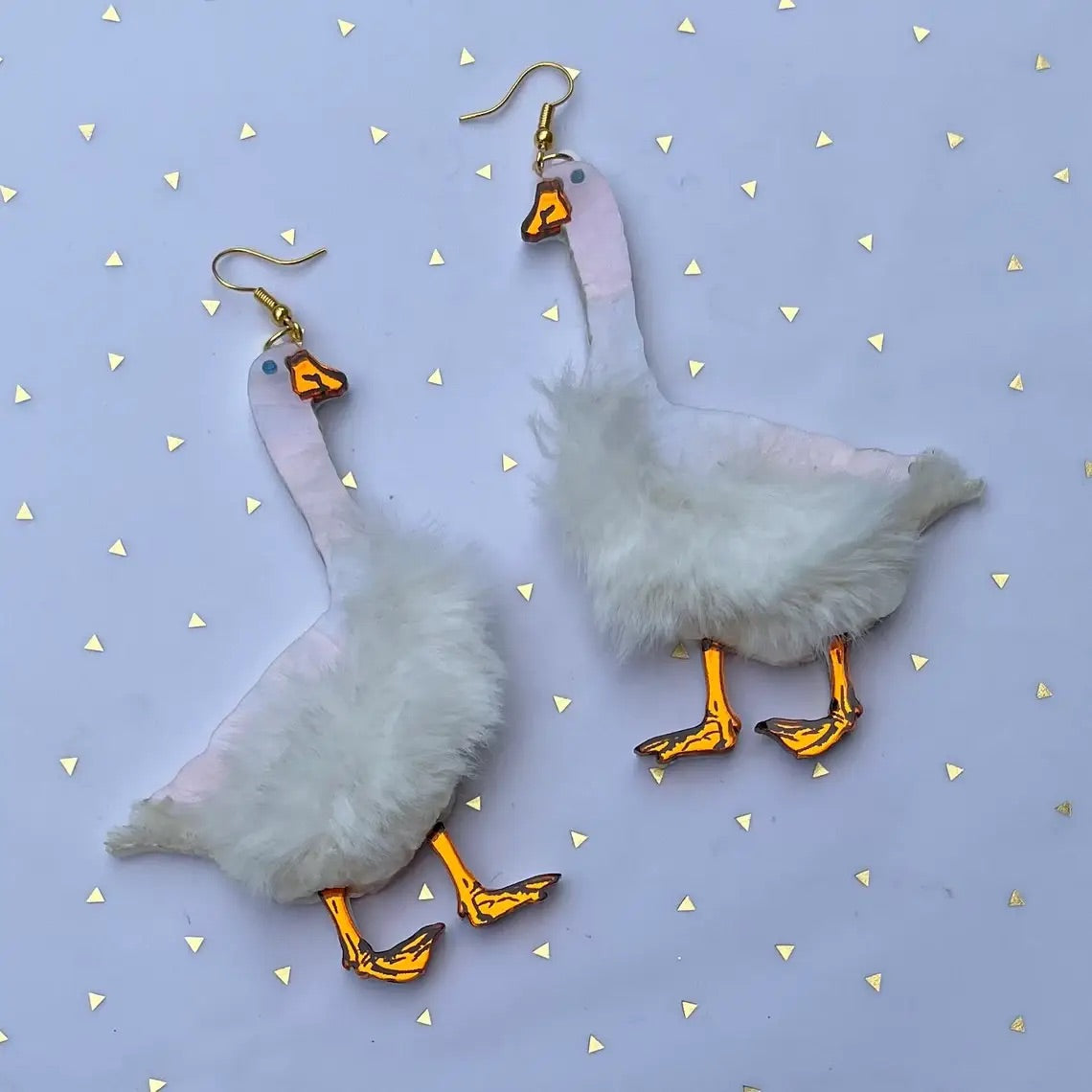 A pair of laser-cut acrylic dangle earrings in the shape of a pair of geese with pearly white bodies, mirrored orange beaks and feet, and small blue eyes. There is a patch of white faux fur on each body. Shown lying flat.