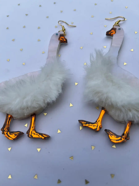 A pair of laser-cut acrylic dangle earrings in the shape of a pair of geese with pearly white bodies, mirrored orange beaks and feet, and small blue eyes. There is a patch of white faux fur on each body. Shown in close up.