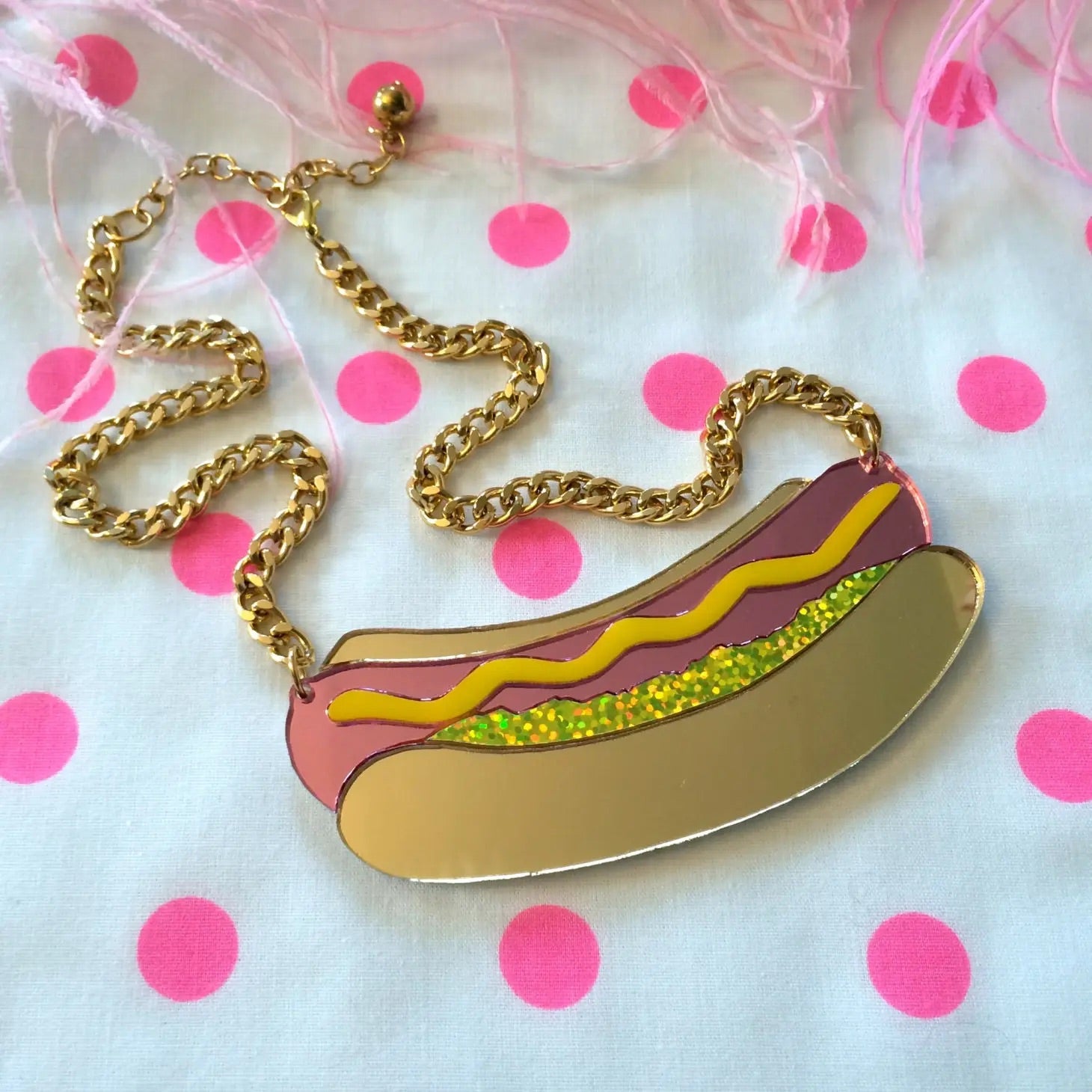 Necklace with large hot dog shaped charm made  of laser-cut layered mirrored acrylic pink hot dog and brown bun with squiggly yellow mustard and green glitter foil relish on a gold metal linked chain