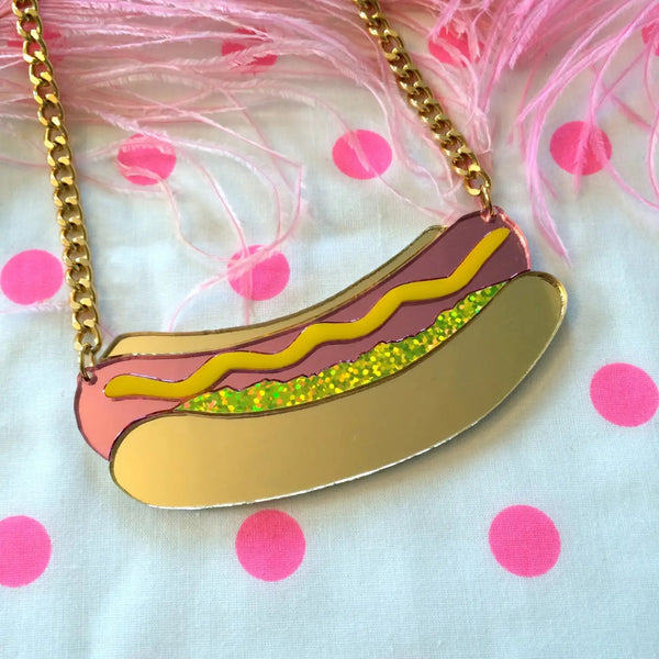 Necklace with large hot dog shaped charm made of laser-cut layered mirrored acrylic pink hot dog and brown bun with squiggly yellow mustard and green glitter foil relish on a gold metal linked chain. Shown in close up
