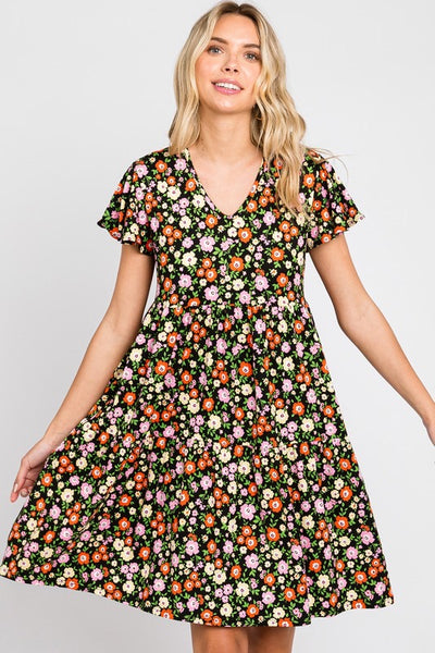 Knit dress in a red, pink, yellow, and green floral pattern on a black background. The dress has a shallow v-neck and short flutter sleeves with a gathered at the waist tiered skirt that ends just above the knee. Shown on a model holding the skirt of the dress wide 