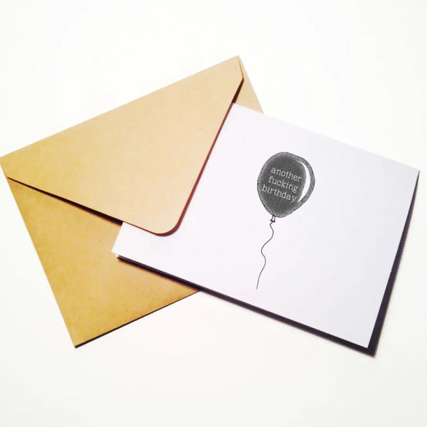 A greeting card with an image of a single black balloon on a string with the message “another fucking birthday”. Shown with included brown envelope 
