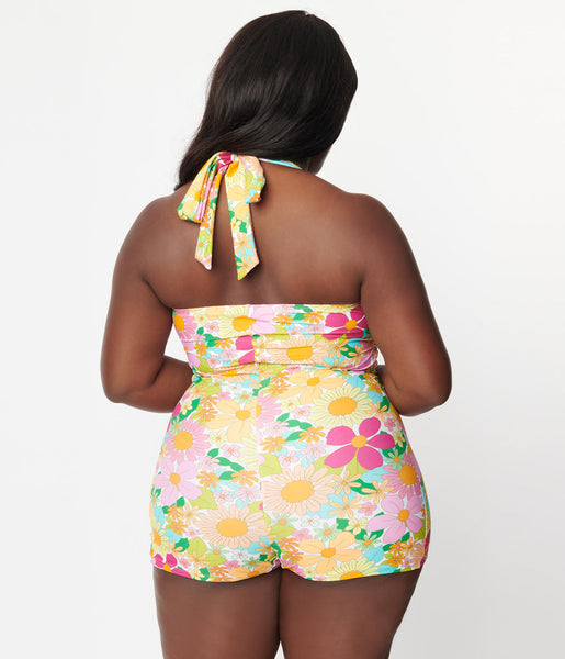 A plus size model model wearing a 50s style halter one piece. It is in a bright pink, orange, yellow, and green 70s style floral pattern. It has wide matching halter straps, a keyhole and tie detail at the bust, slightly ruched waist, and boy short style bottom. Shown from behind to illustrate halter tie detail