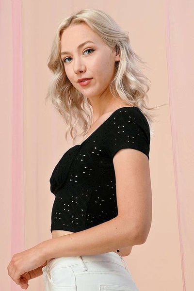 Black eyelet fabric crop top with square neckline, a lightly ruched bodice, and cap sleeves. Shown on a model from the side