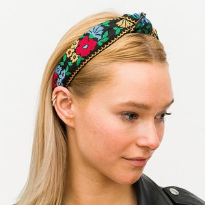 2” wide embroidered fabric headband in a red, blue, yellow, and green floral fabric pattern on a black background with yellow scalloped detail at the edges. Worn by a model