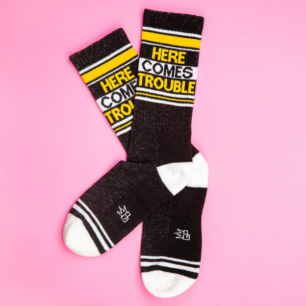 "Here Comes Trouble” text yellow, black, and white ribbed knit stretch cotton blend crew length gym socks, shown flat on a pink background 