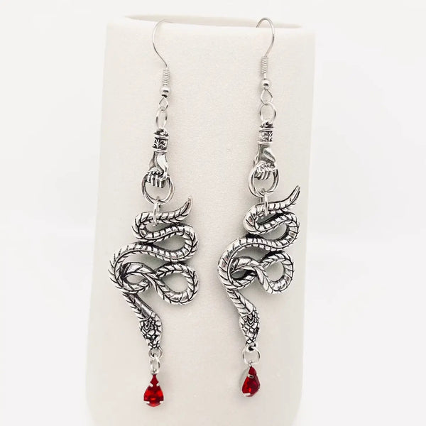 silver metal dangle earrings featuring a hand-shaped charm holding a stylized snake with a bright red faceted bead in the shape of a drop of blood