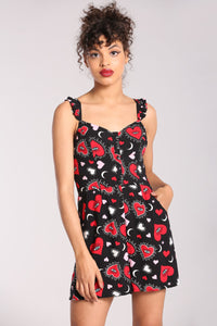 A model wearing a playsuit in a red, pink, and white heart, star, and eye pattern on a black background. It has spaghetti straps with ruffle detail, buttons down the sweetheart bodice, and high-waisted wide legged shorts with pockets. Seen from the front 