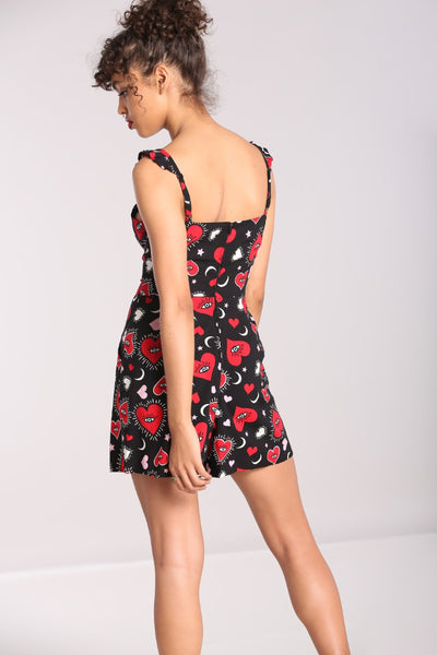 A model wearing a playsuit in a red, pink, and white heart, star, and eye pattern on a black background. It has spaghetti straps with ruffle detail, buttons down the sweetheart bodice, and high-waisted wide legged shorts with pockets. Seen from the back 