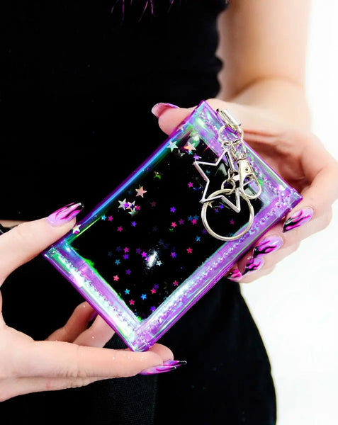 Rectangular wallet made of silver and iridescent purple vinyl filled with liquid glitter in purple, blue, black, and silver holographic star shaped glitter. It has a silver metal star shaped key ring. Shown in close up