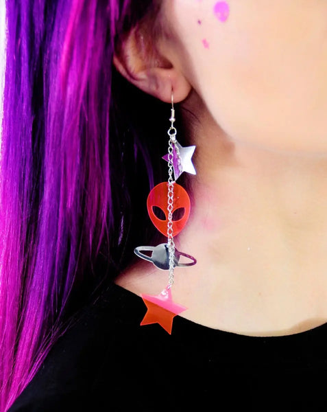 A pair of dangle earrings made of pieces of neon pink and silver chrome vinyl in alien, star, and planet shapes connected by pieces of silver metal chain. Shown on the ear of a model  