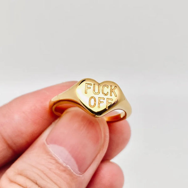Gold plated stainless steel ring shaped like a heart with “FUCK OFF” etched onto the top. Shown held up by a hand showing front 