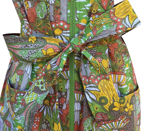 A short sleeved mini dress with an exaggerated pointed collar and green plastic zipper down the front. It has a psychedelic pattern with motifs including star spotted mushrooms, daffodils, paisley, snails, and pinwheels- in shades of yellow, red, green, and black. Shown in close up zipped with sash belt