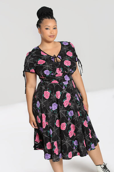 Plus-size model wearing a mid length rayon dress in a stylized white spiderweb with purple and pink rose pattern on a black background. It has keyhole and tie details at both the slight v-neckline and the short sleeves. The bodice is slightly gathered under the bust and the skirt is flared just below the knee. Shown from the front 