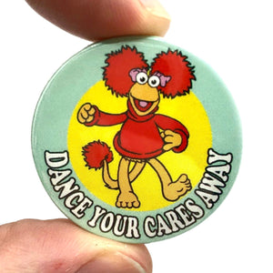 1 1/2” round pinback button with the caption “DANCE YOUR CARES AWAY” below an illustration of a character from the tv show Fraggle Rock