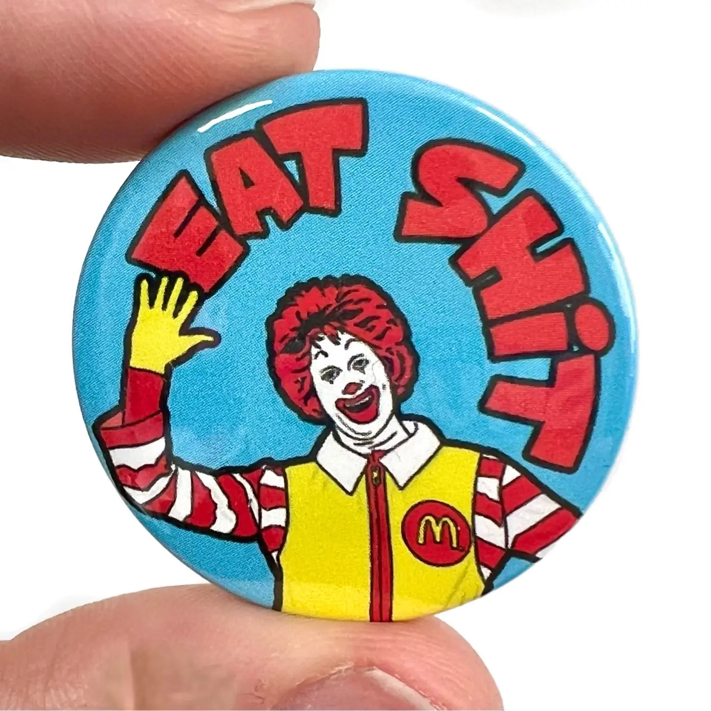 1 1/2” pinback button with the words “EAT SHIT”  in red above a cartoon clown mascot with a light blue background 