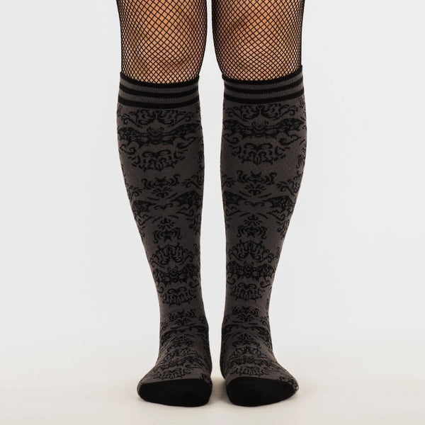 Knee socks with a monochromatic damask pattern of flying bats. Striped grey and black bands at the top of each sock and solid black toes and heels. Shown on a model wearing fishnet tights underneath the socks, shown from the front